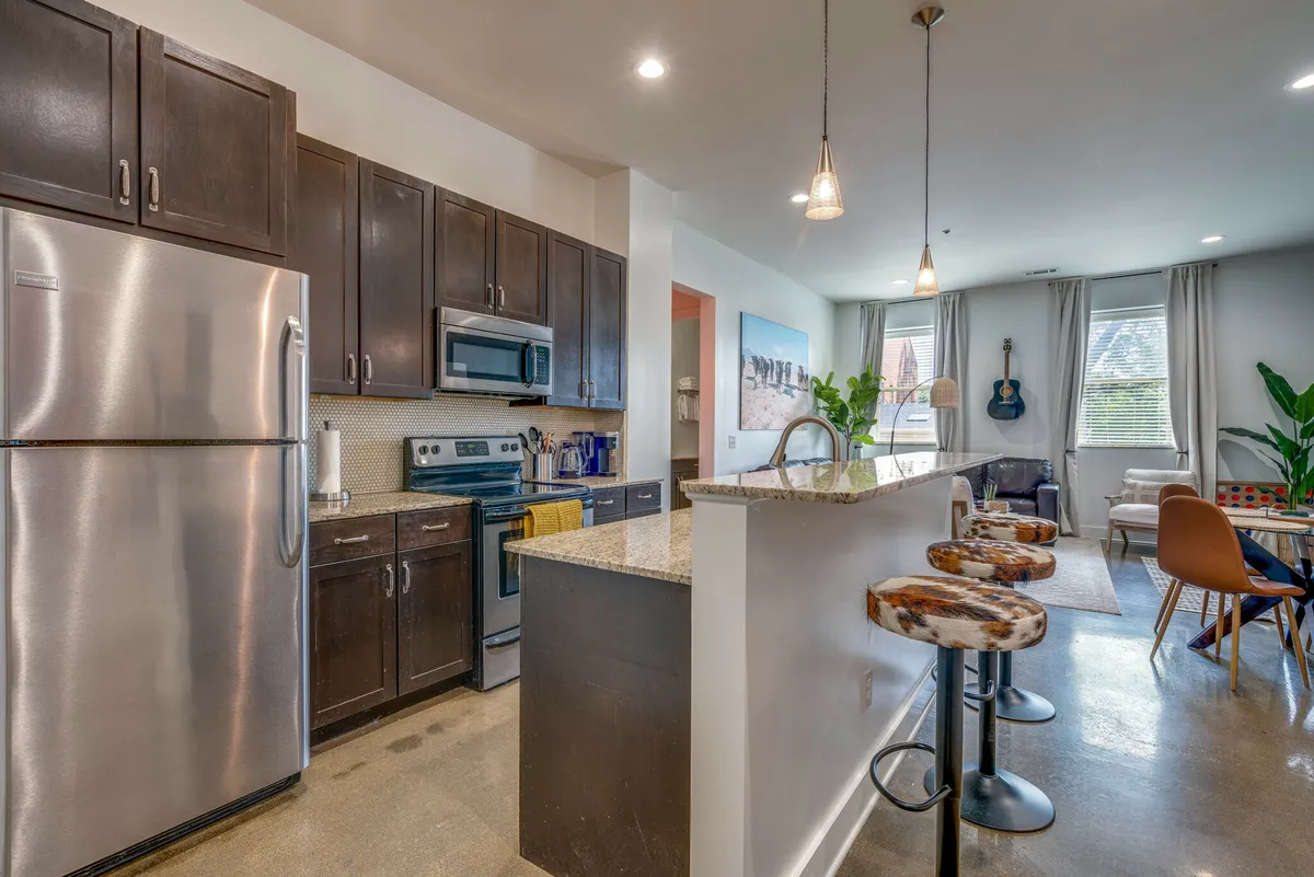 A welcoming and relaxed kitchen space is highlighted by gleaming stainless steel appliances, a wide granite countertop with bar stools, and lively decor including a guitar on the wall and indoor plants. The lighting and layout invite both cooking and socializing, offering ample room for meal preparation and casual gatherings.