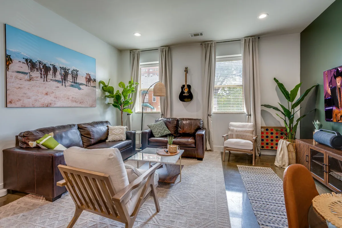 The image shows a cozy and stylish living room with large, playful cow artwork, a guitar hanging on the wall, lush green plants, and a comfortable seating area centered around a TV—perfect for relaxing or socializing.
