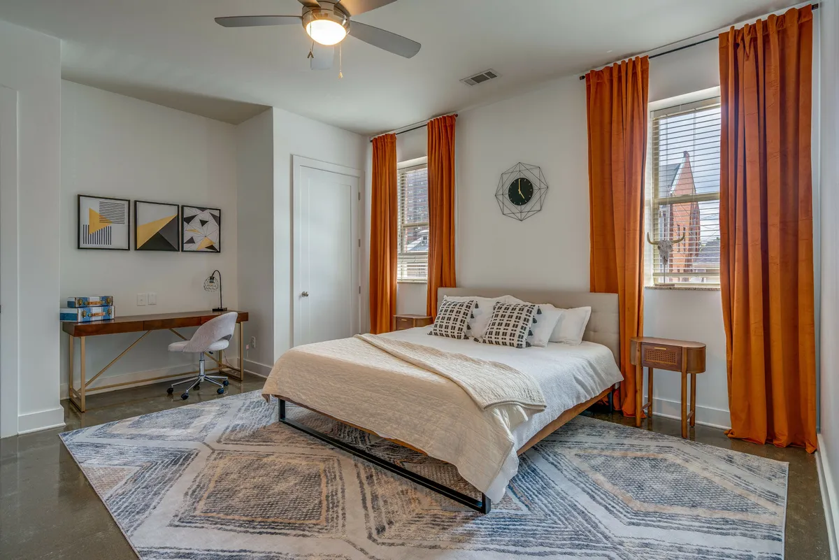 A bright, tranquil bedroom featuring a large bed with orange curtains framing sunlight streaming into the room. There's a cozy rug on the floor and a small desk positioned for work or reading, creating a space that balances relaxation and productivity.