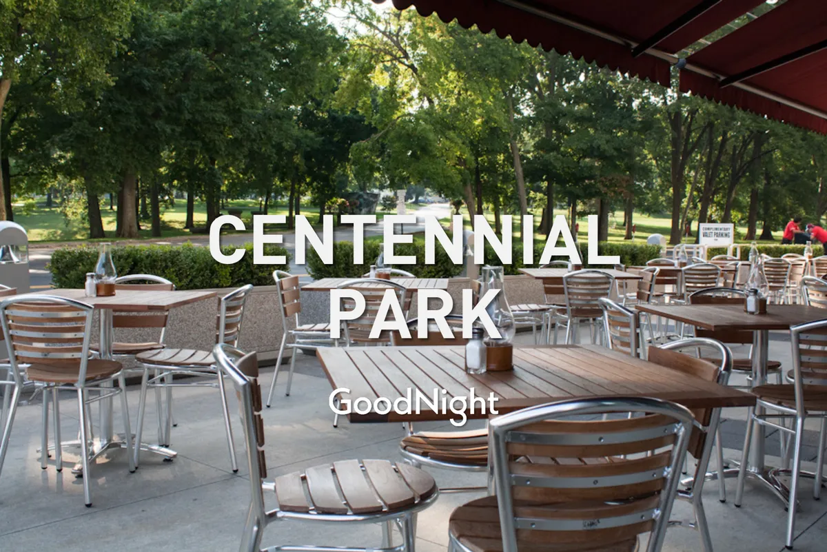This image captures an exterior park scene with wooden tables and metal chairs set amidst green foliage by a park pathway. The area looks perfect for outdoor dining, casual gatherings, and picnics, and the visible trees provide ample shade.