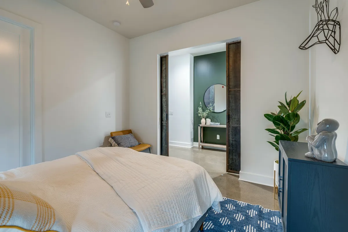 A calm and inviting bedroom with a plush bed against a green accent wall, a decorative mirror, a leafy plant, and a unique sculpture. The space feels cozy, modern, and ideal for peaceful moments with loved ones.