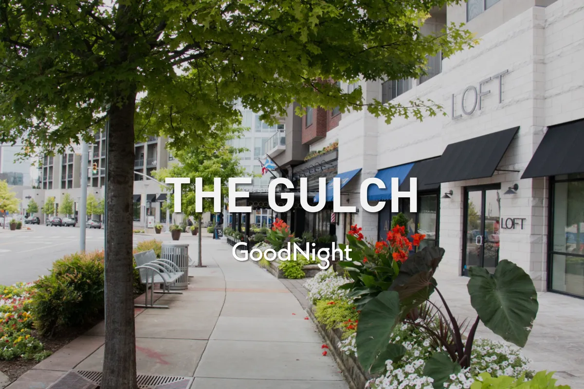This image shows a vibrant, tree-lined sidewalk bordered by blooming flower beds, storefronts, and benches. The scene is lively, with clear evidence of walkability, outdoor leisure, and local dining or shopping options.